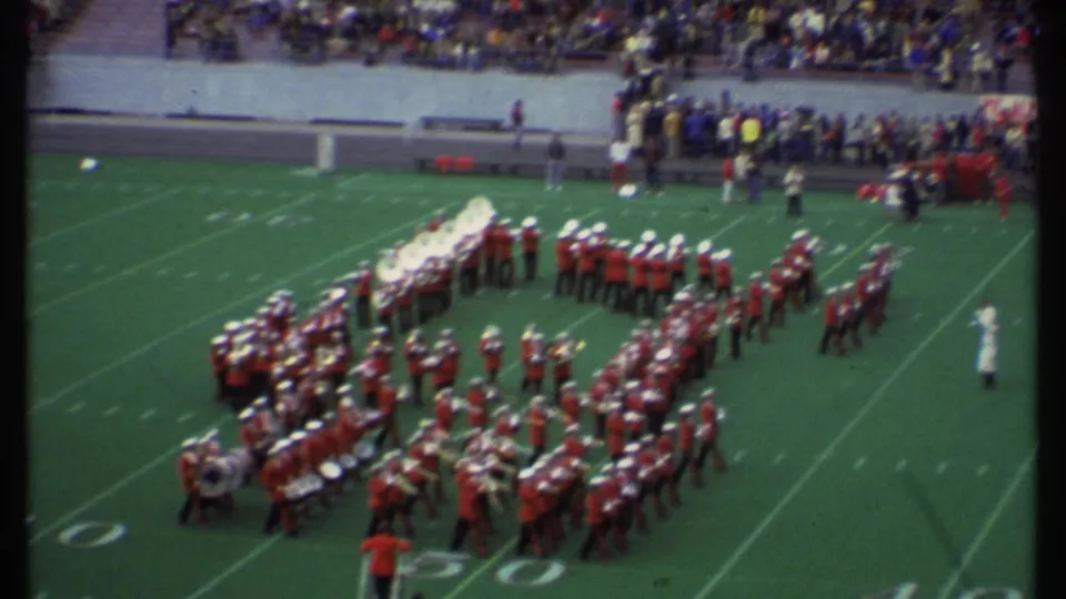 Red-and-white marching band continues a formation on the Ithaca football field, filmed from an elevated spectator position in 1979.