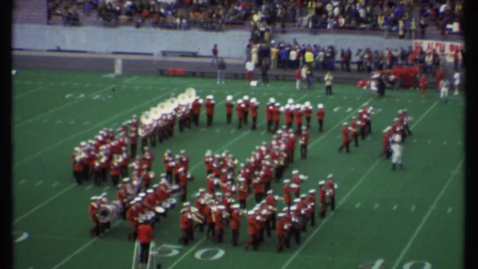 8mm home-movie view of a marching band in red uniforms shifting formation on a football field in Ithaca, New York in 1979.