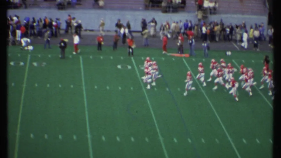 Players in red and white uniforms run onto the football field in Ithaca, New York after the marching band segment, circa 1979.