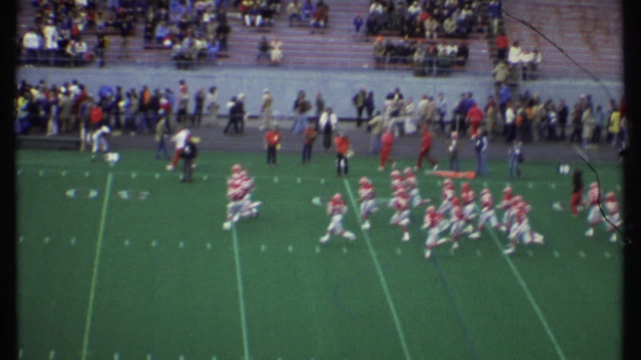 Football players in red and white uniforms run across a green field with spectators in bleachers behind them.