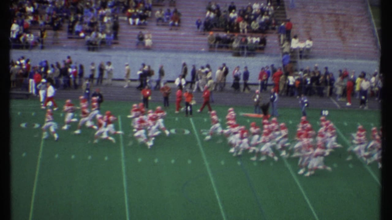 Two football teams move during a play on a marked field with a crowd seated and standing in the background.