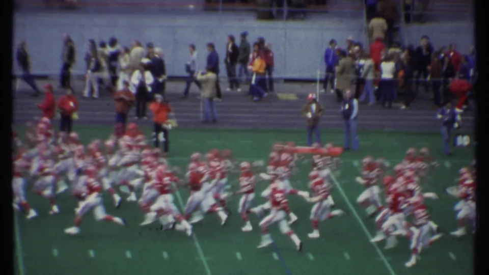 Late-1970s 8mm football frame from Ithaca, New York, showing red and white players moving past a crowded sideline.