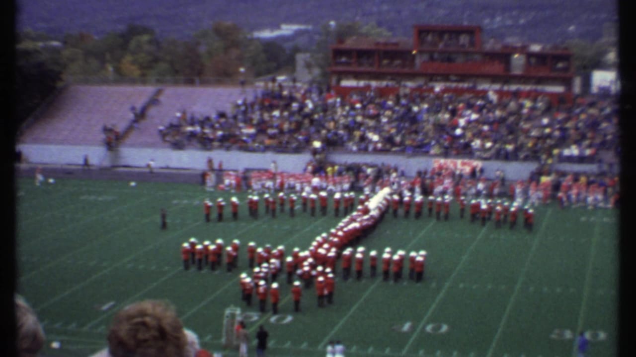 Marching band in red uniforms forms a large letter on a football field before crowded bleachers.