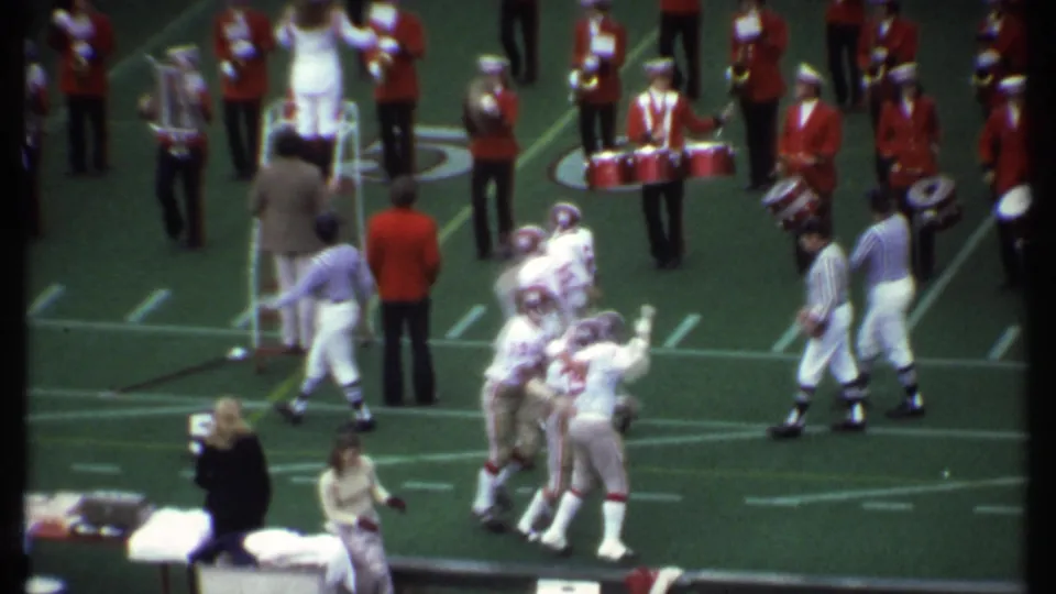 Football players in white uniforms celebrate on an Ithaca field while red-uniformed band members and sideline figures stand nearby.