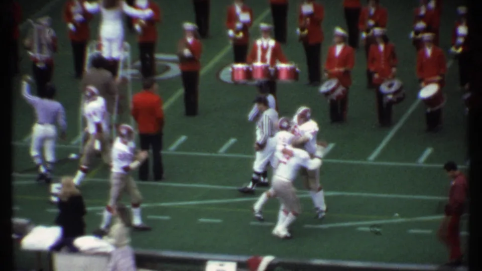 Players in white football uniforms embrace and walk across the Ithaca field, with red-uniformed band members visible behind them.