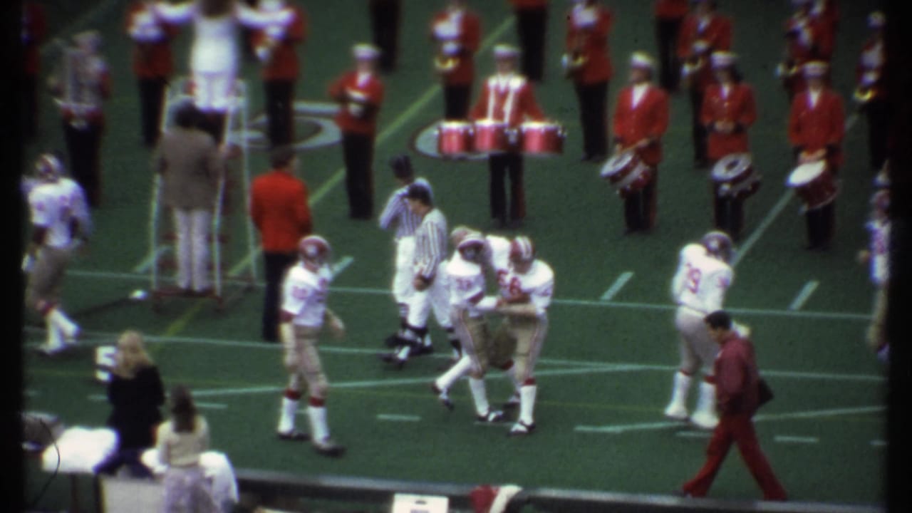 Football players gather near the sideline while a marching band stands behind them on the field.