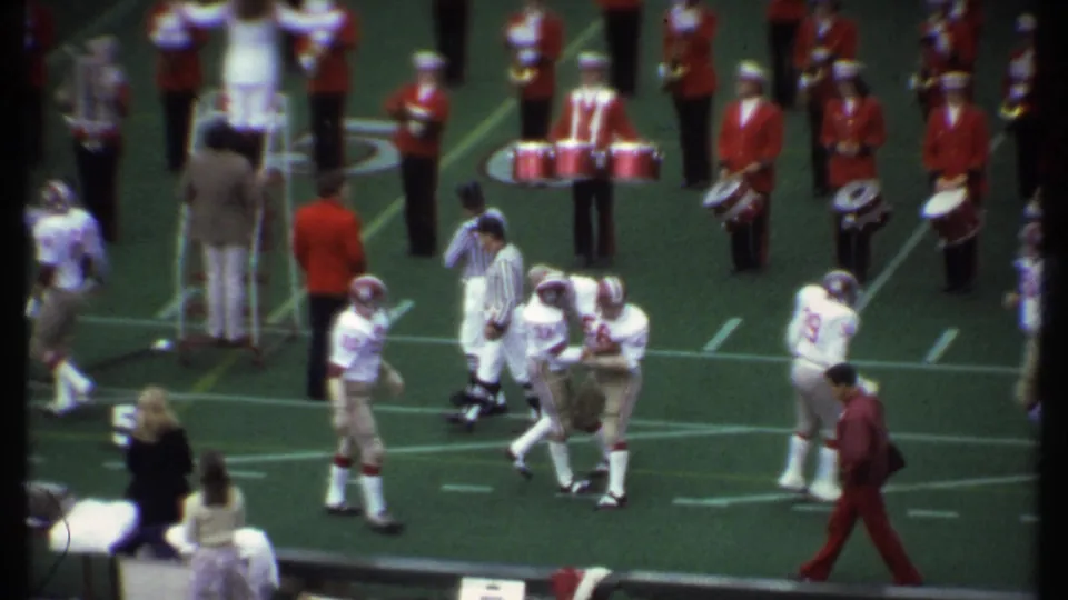 In Ithaca, New York, 1979, football players in white and gold uniforms move near the sideline as a red-uniformed marching band waits behind them.
