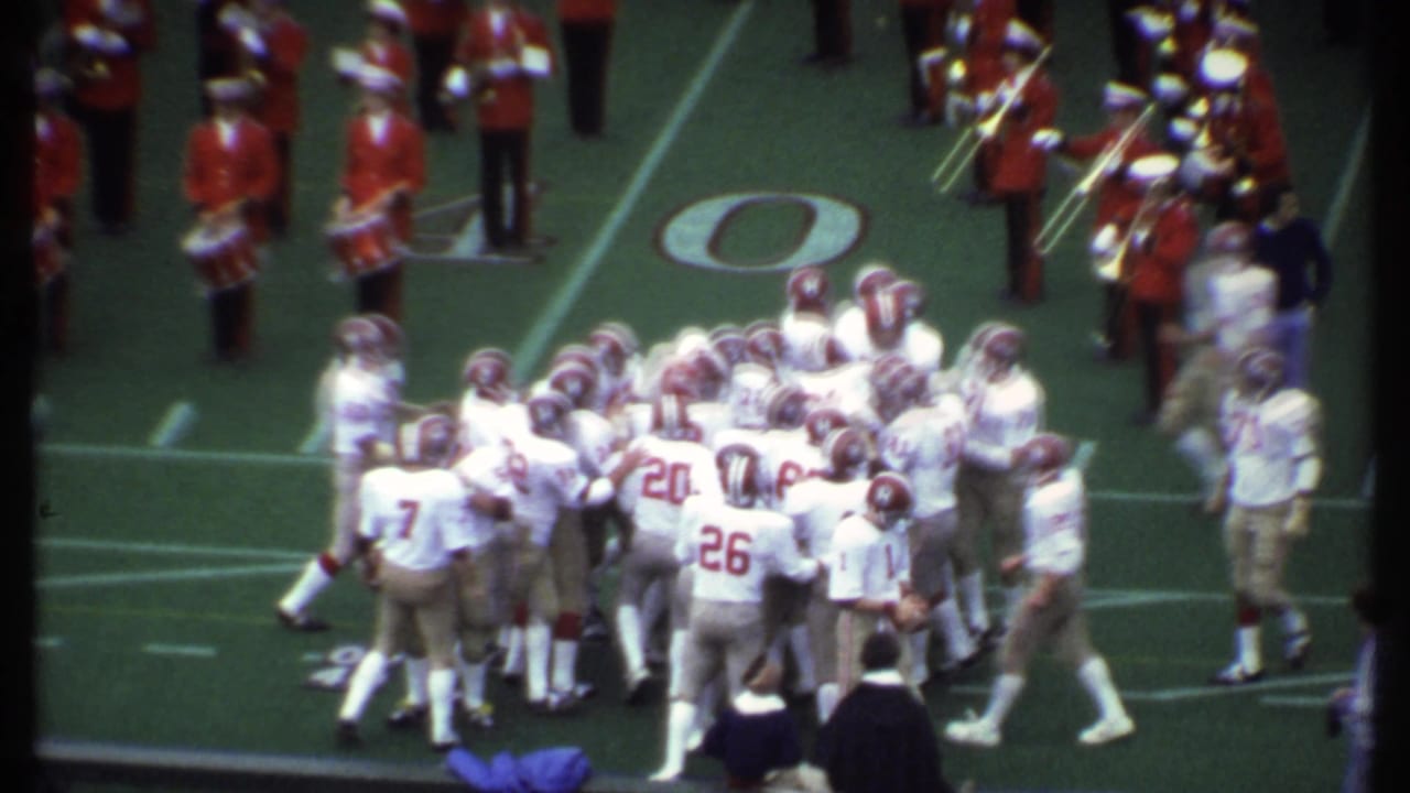 Football players in white uniforms huddle on the field with band members visible behind them.