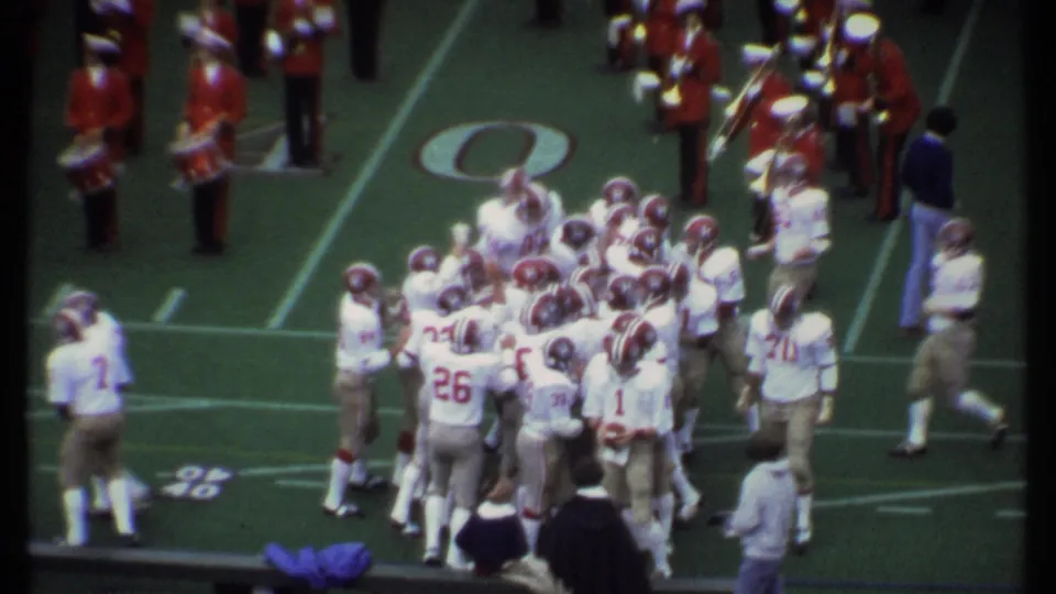 Football players cluster in a huddle during the 1979 Ithaca game, with marching band members and sideline figures visible around the field.