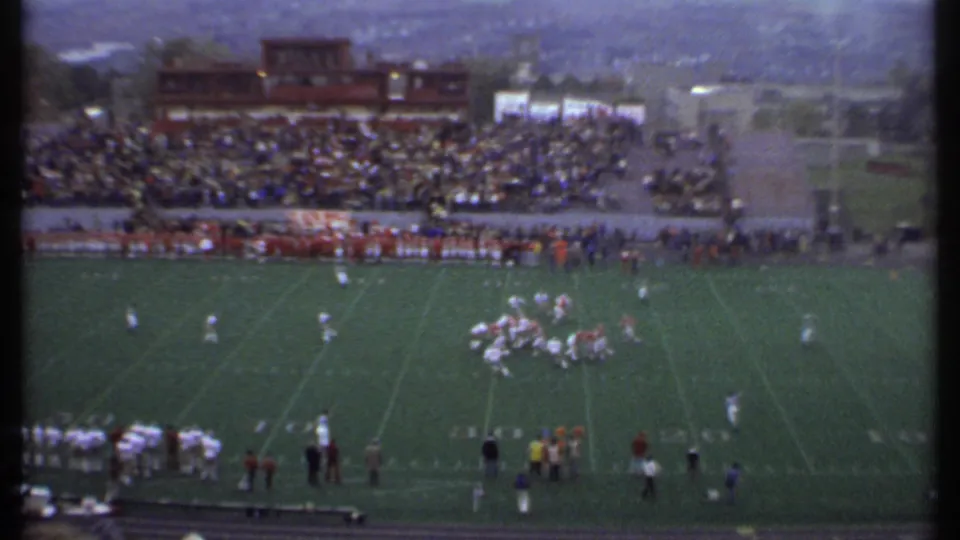 Wide 1979 Ithaca stadium view showing football players lined up on the field, spectators in the stands, and sideline groups along the lower edge.