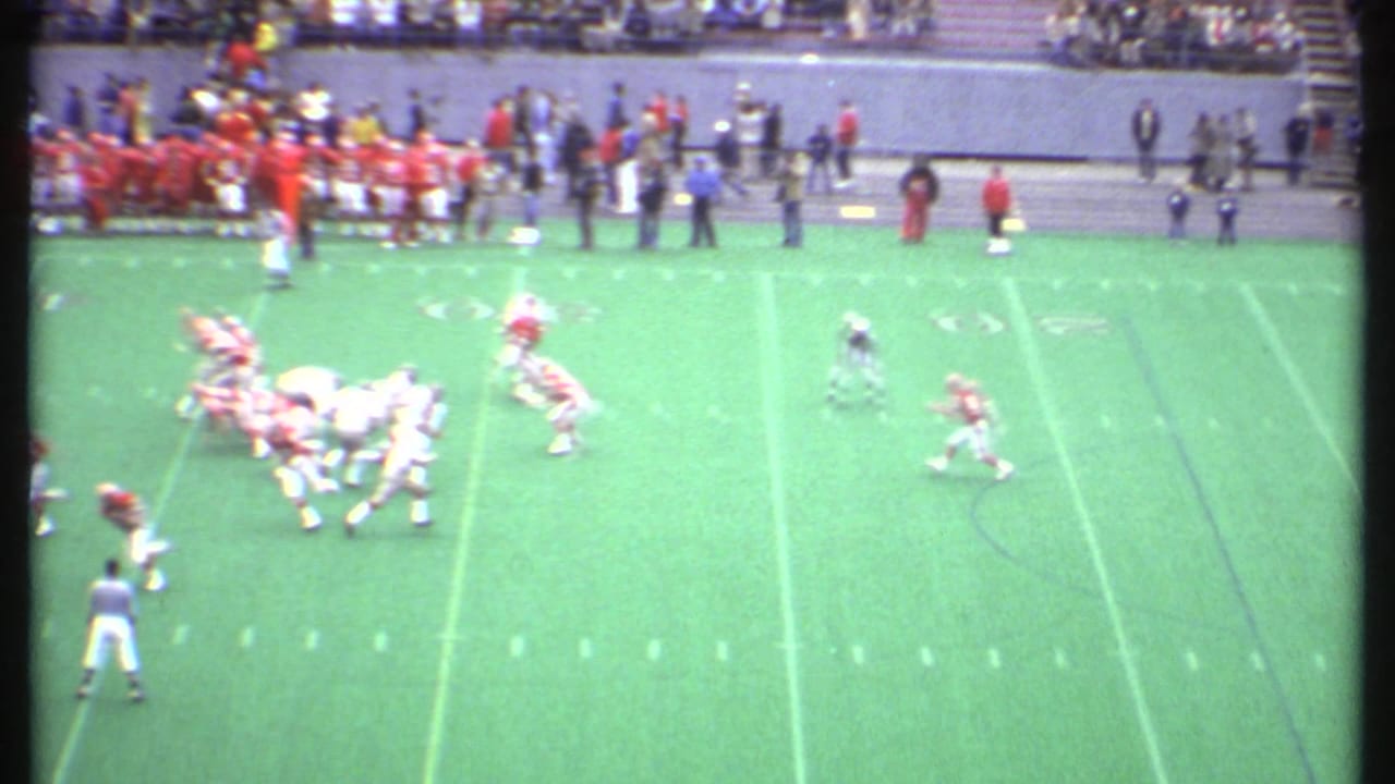 Football teams line up on a green field with spectators and sideline figures in the background.