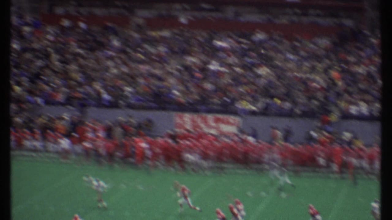 A football play moves downfield with a crowded stadium and red-uniformed sideline behind it.