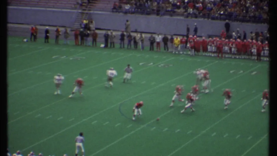 A wide 8mm stadium view shows red and white football players moving through a play on the marked field.