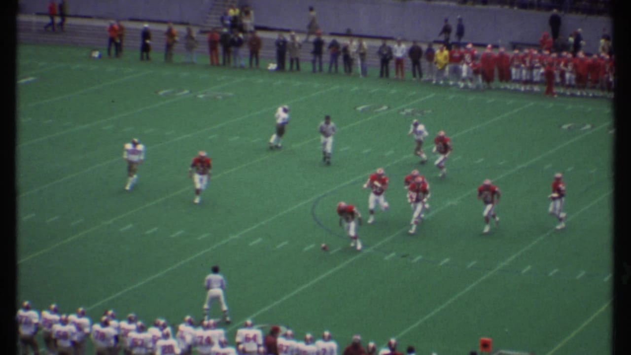 Football players line up on a green field with sidelines and spectators visible in the distance.