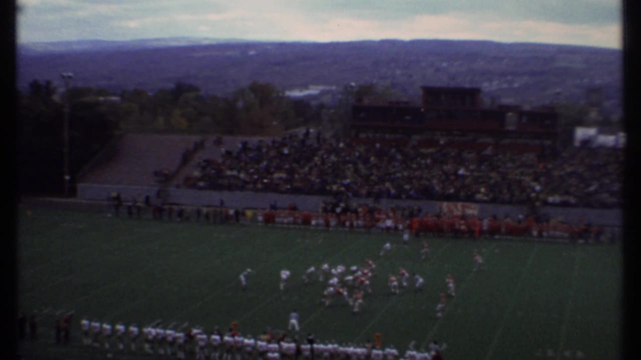 Wide stadium view with a football play underway, packed bleachers, hills, and cloudy sky beyond.