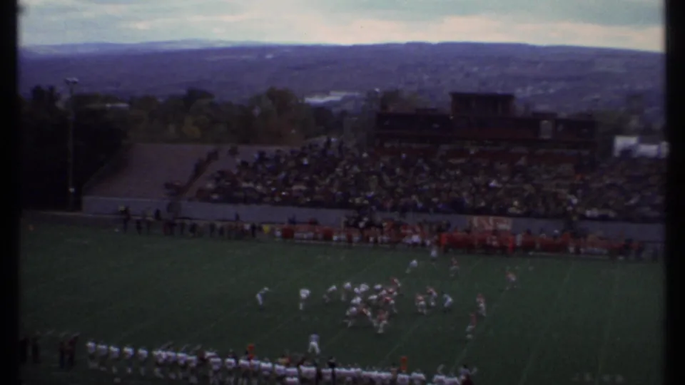 Wide 8mm view of a 1979 Ithaca football game with the play on the field below crowded bleachers and distant hills.