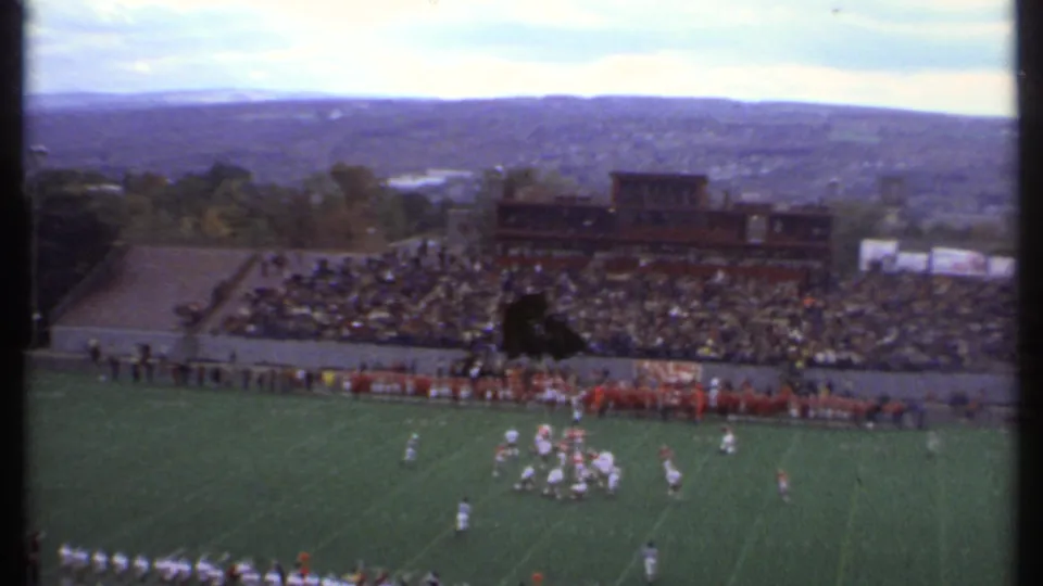 1979 Ithaca football footage showing players regrouping on the field, with a full stadium section and wooded hills beyond.