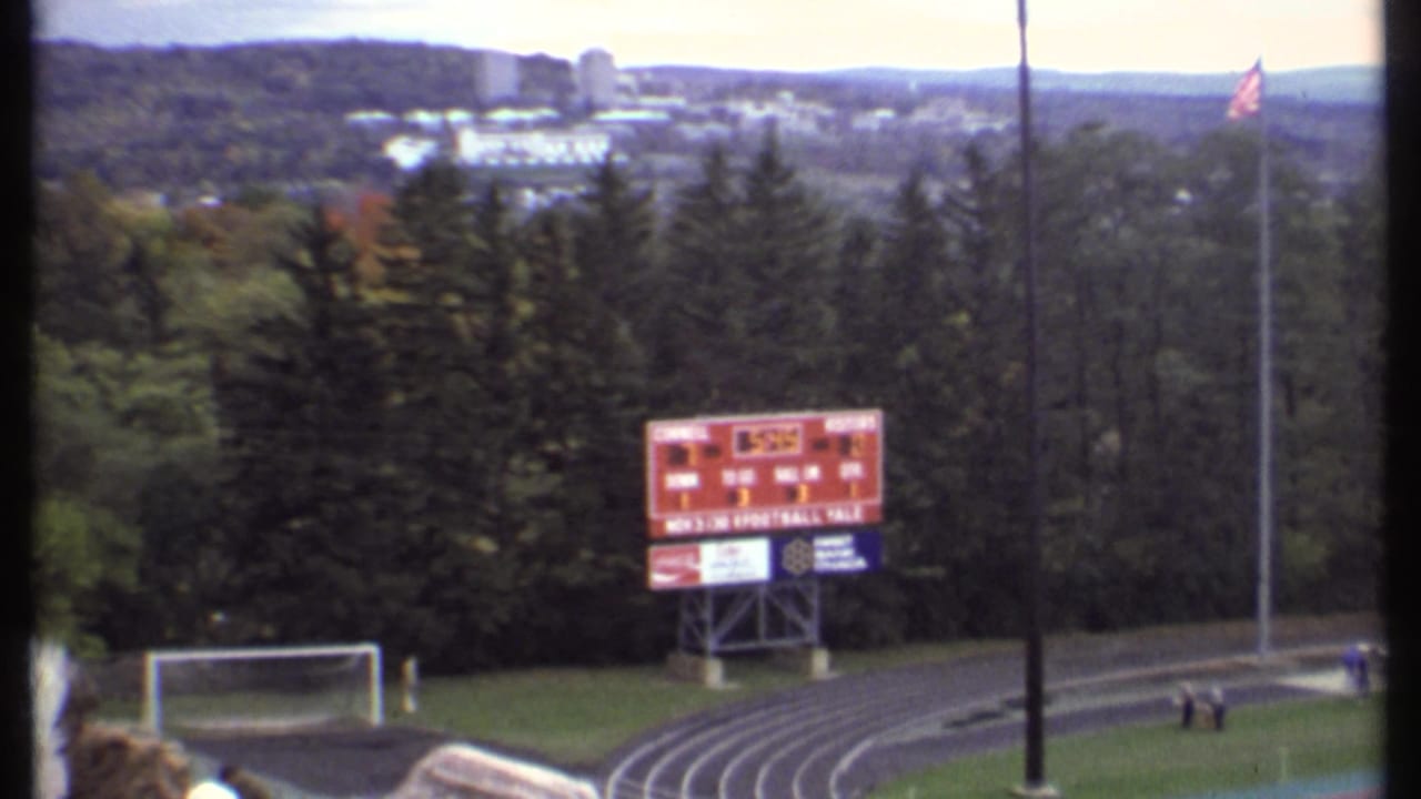 Wide stadium view with football field, track, scoreboard, trees, and flags.