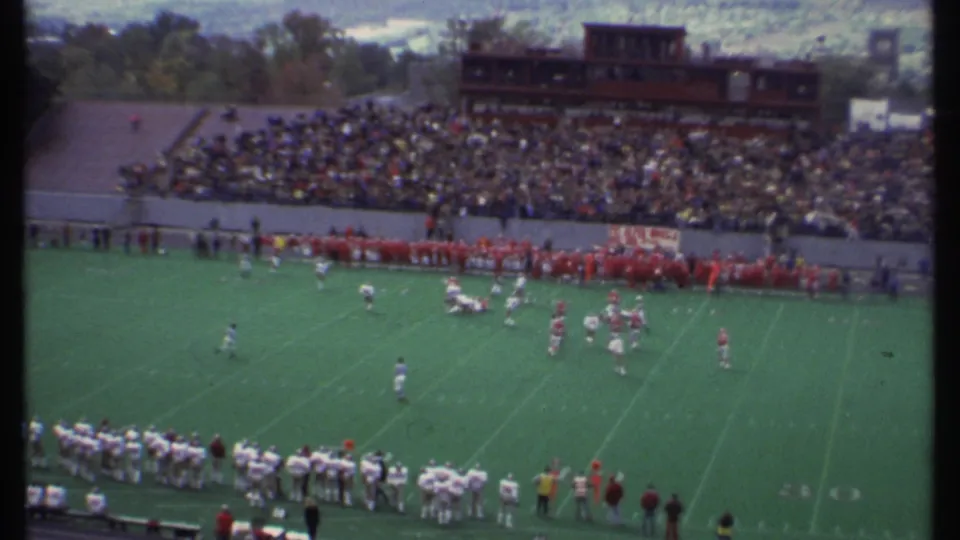 1979 Ithaca football action from a high spectator angle, showing players on the field, sideline groups, and a dense crowd.