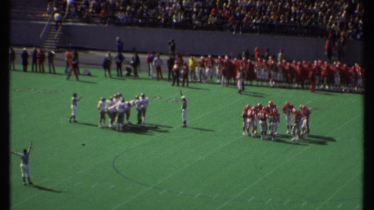 Football teams in red and white huddle on a green field before a packed stadium crowd.