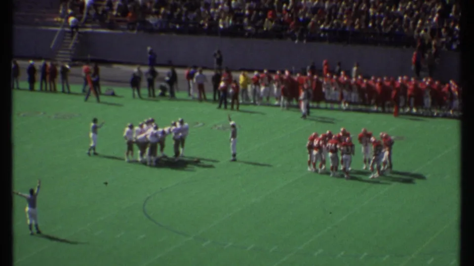 Distant 1979 Ithaca football game view with red and white teams huddling on the field and spectators filling the stands.