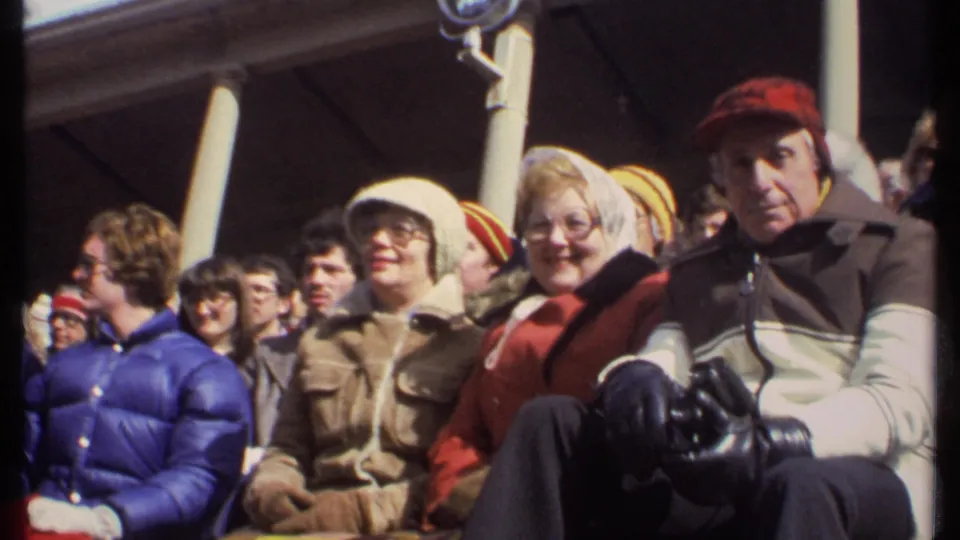 Adults in winter clothing sit in the Ithaca stadium crowd, with several faces turned toward the home-movie camera.