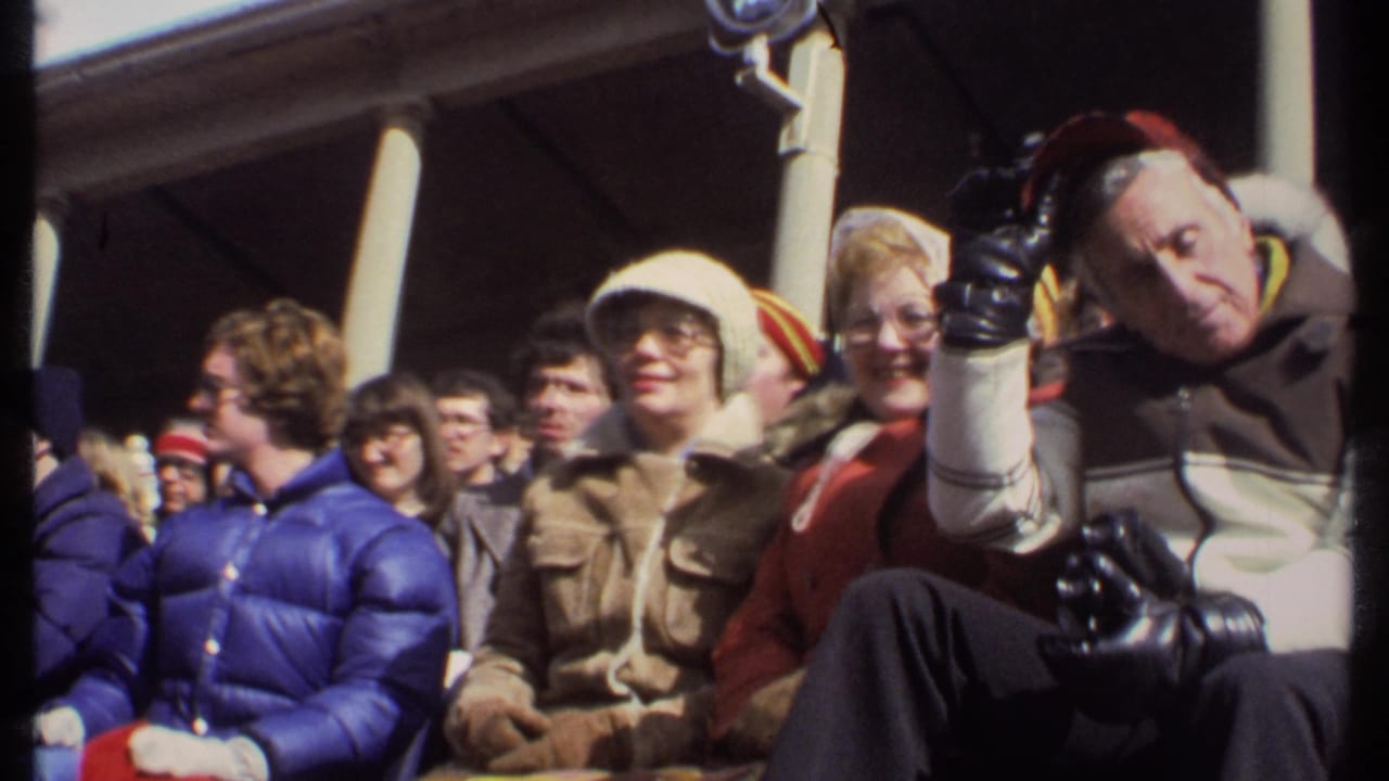 1979 Ithaca home-movie frame of football spectators in winter coats, including a man tipping his red cap toward the camera.