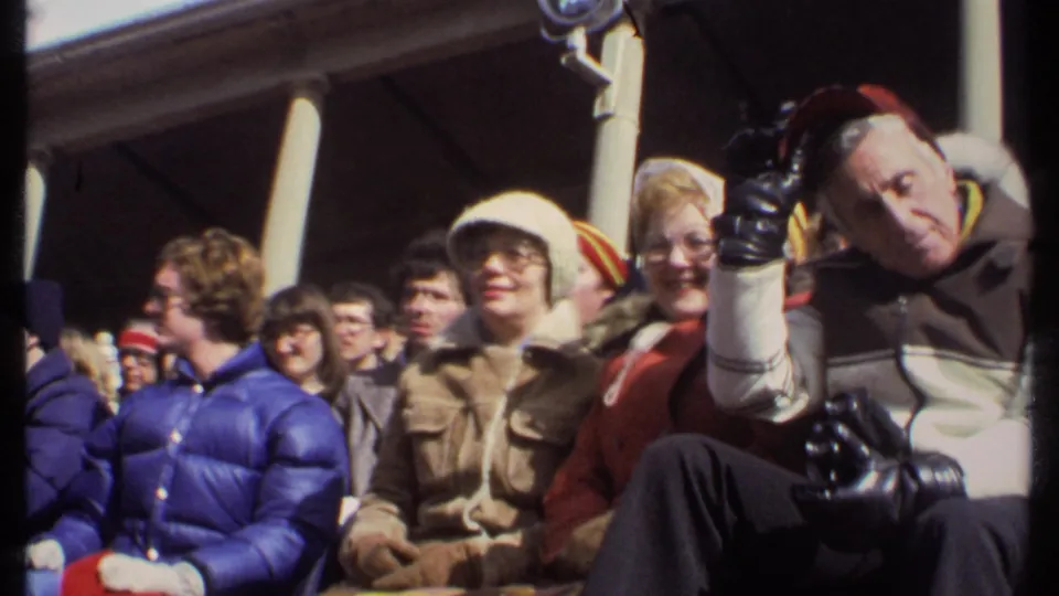 1979 Ithaca home-movie frame of football spectators in winter coats, including a man tipping his red cap toward the camera.