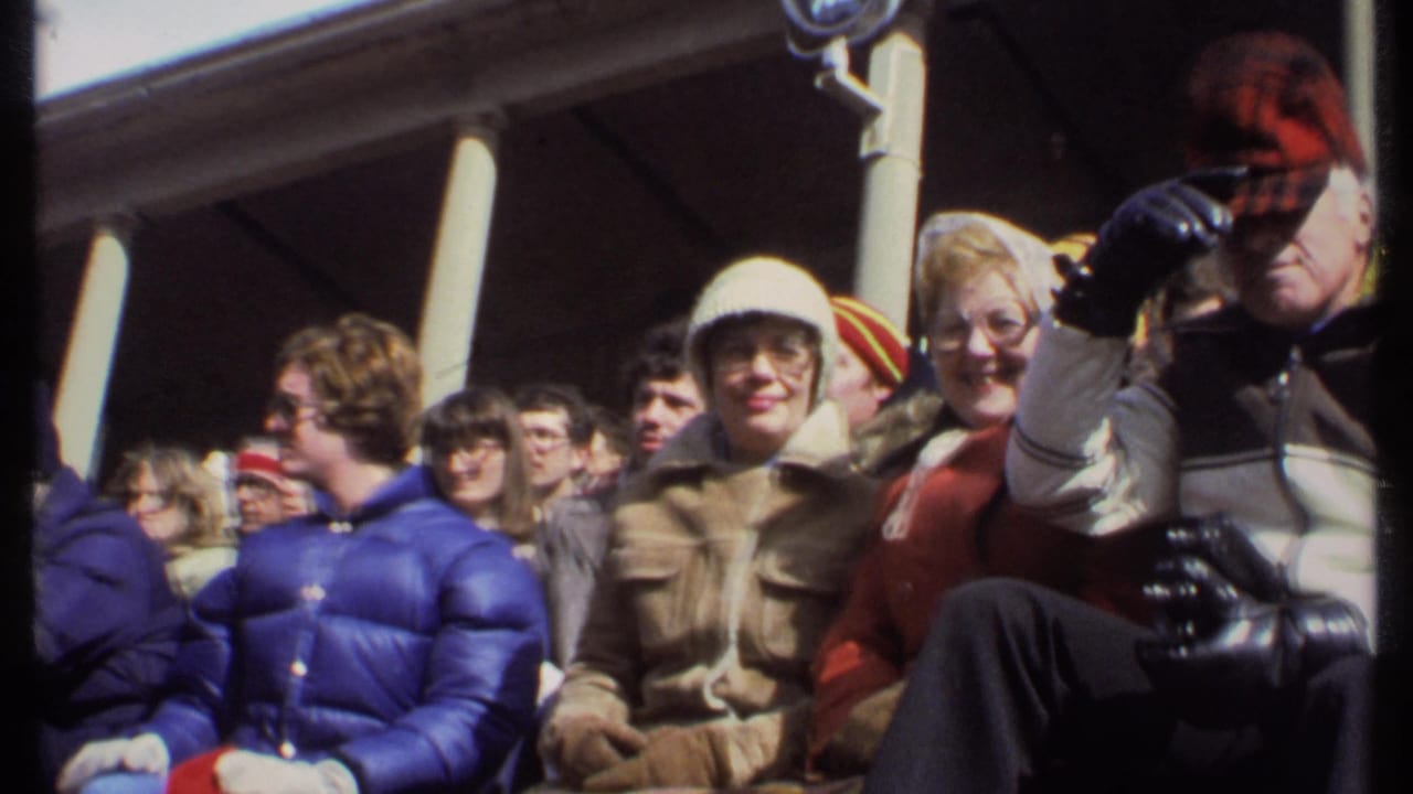 Crowd seated in stadium stands as a man in a red cap tips his hat close to camera.
