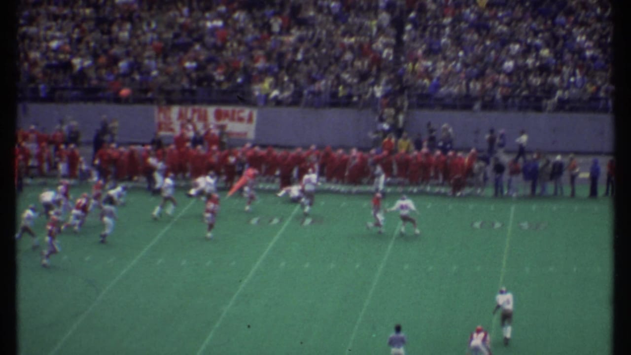 Football players in red and white uniforms run across a marked field before stadium spectators.