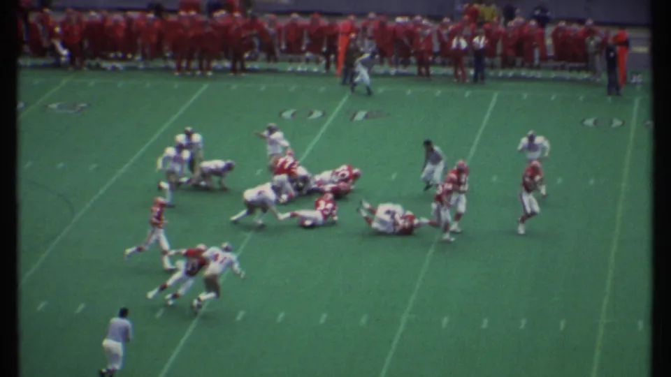 A 1979 Ithaca football play shows players in red and white uniforms going to the ground as a referee and sideline team watch.