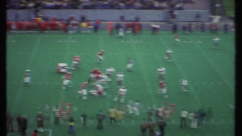 Wide 8mm view of a 1979 Ithaca football game, with red and white players spread across the field and spectators along the far side.