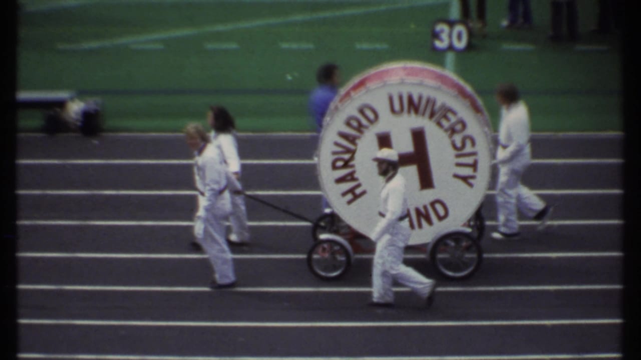 Harvard University Band members wheel a large bass drum along a football sideline.