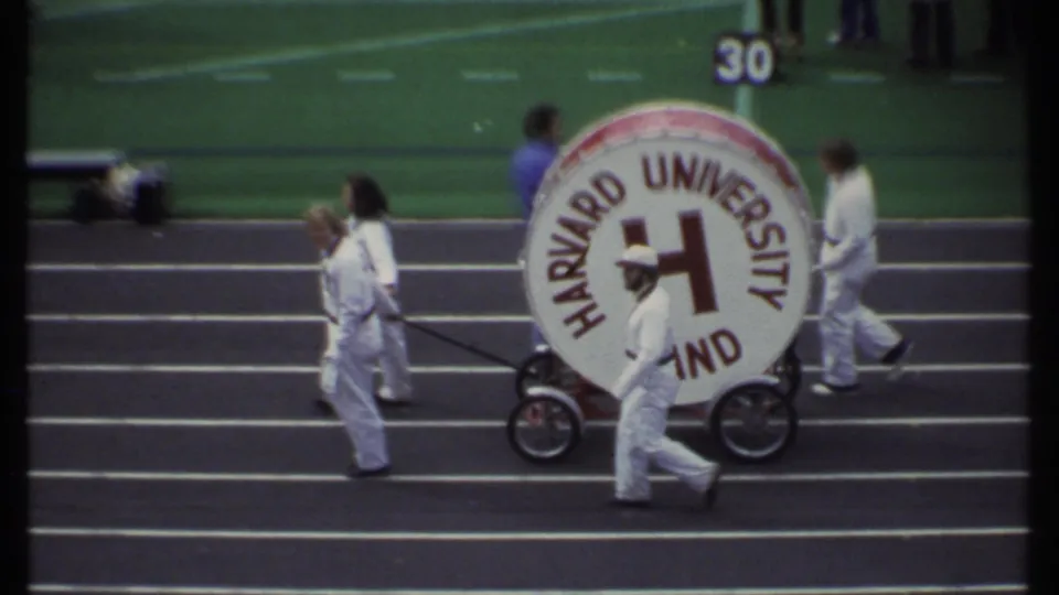 In Ithaca, New York in 1979, white-uniformed Harvard University Band members move a large logo bass drum near the field.