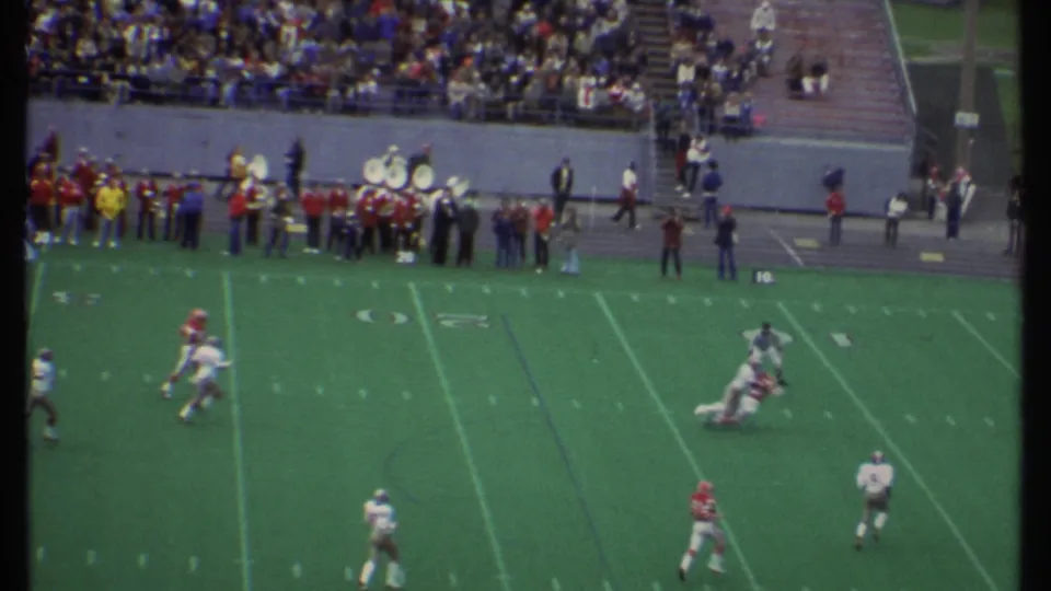 At the Ithaca college football game, players in red and white uniforms move on the field below the band and spectators.