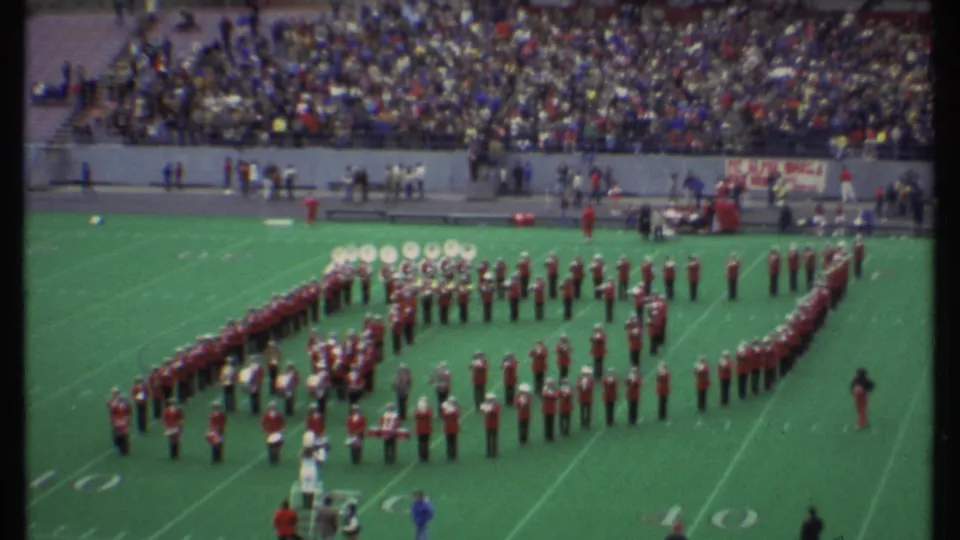 1979 Ithaca, New York home-movie frame showing a red-and-white marching band in formation on a football field with spectators in the stands.
