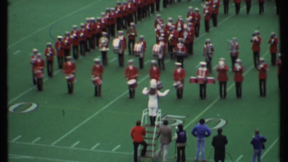 1979 Ithaca, New York frame with a conductor in white directing red-uniformed marching band members on the field.