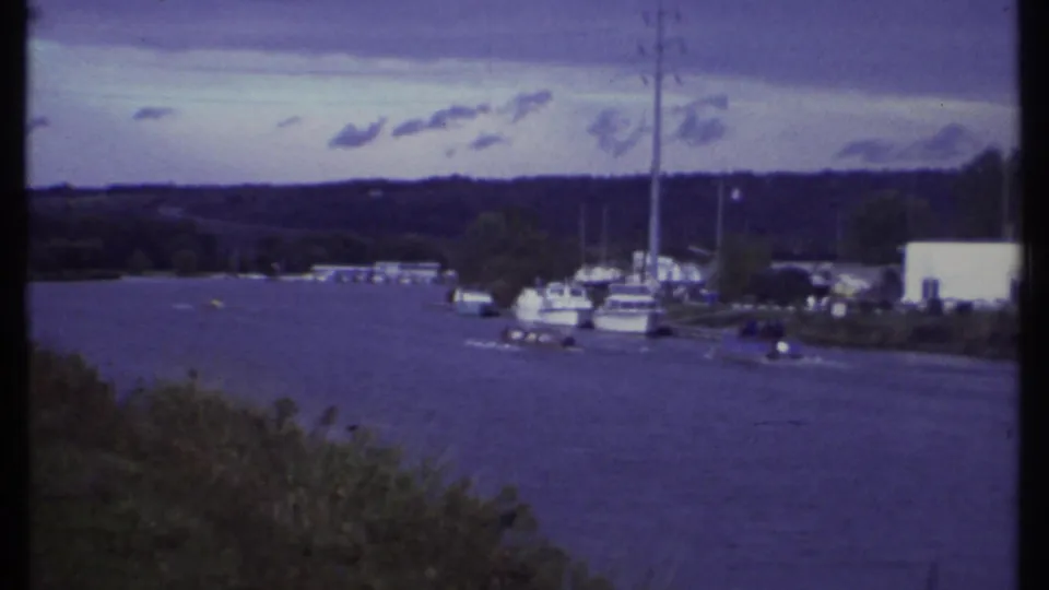 Ithaca, New York waterfront in 1979, with water, marina boats, masts, grassy bank, low buildings, clouds, and wooded shore.