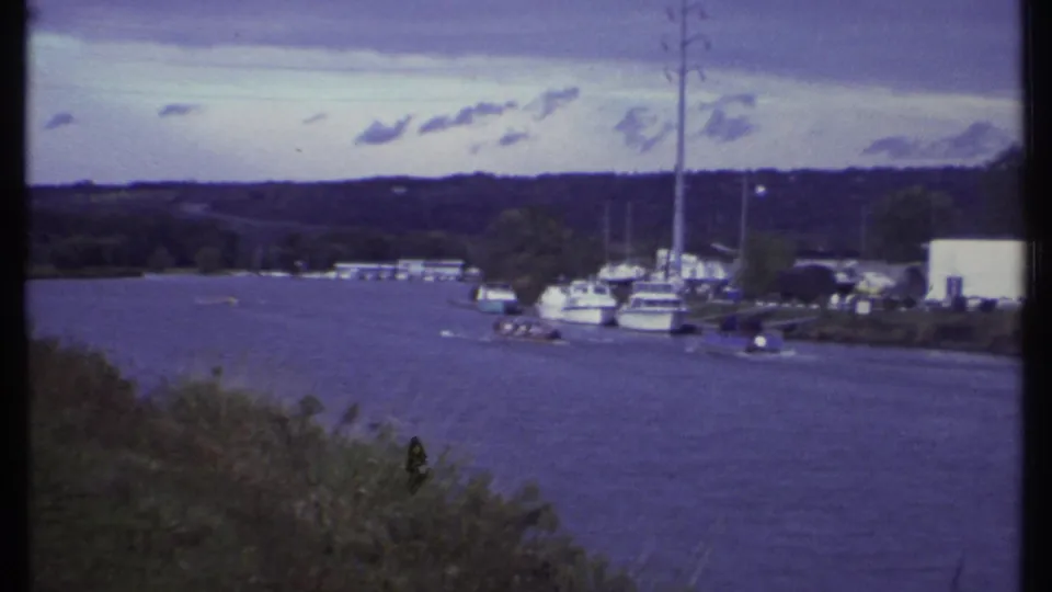 Ithaca, New York 1979 waterfront view showing blue water, distant boats, masts, shoreline trees, low structures, and a grassy foreground.