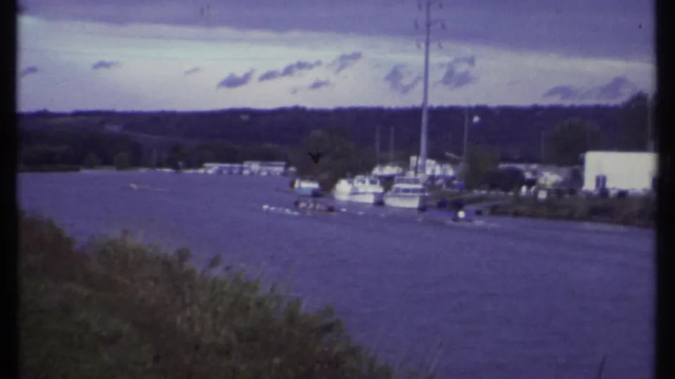 1979 Ithaca home-movie frame showing a lake, marina docks, sailboats, and distant rowers near a wooded shoreline.