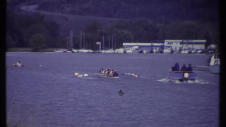 1979 Ithaca lake scene with scullers, a small motorboat carrying passengers, docked sailboats, and a wooded shore.