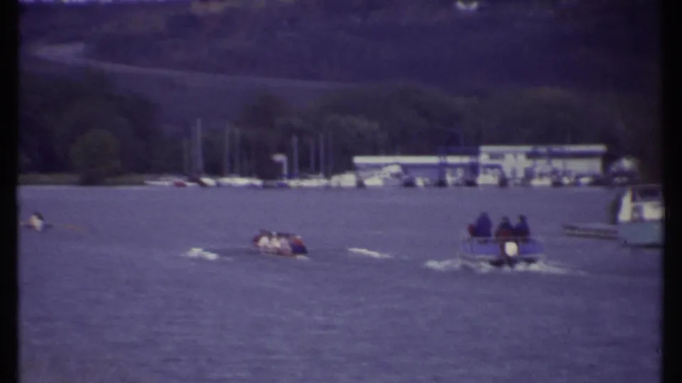 Distant 1979 Ithaca lake footage showing rowers, a passenger motorboat, sailboat masts, and a marina along the shore.