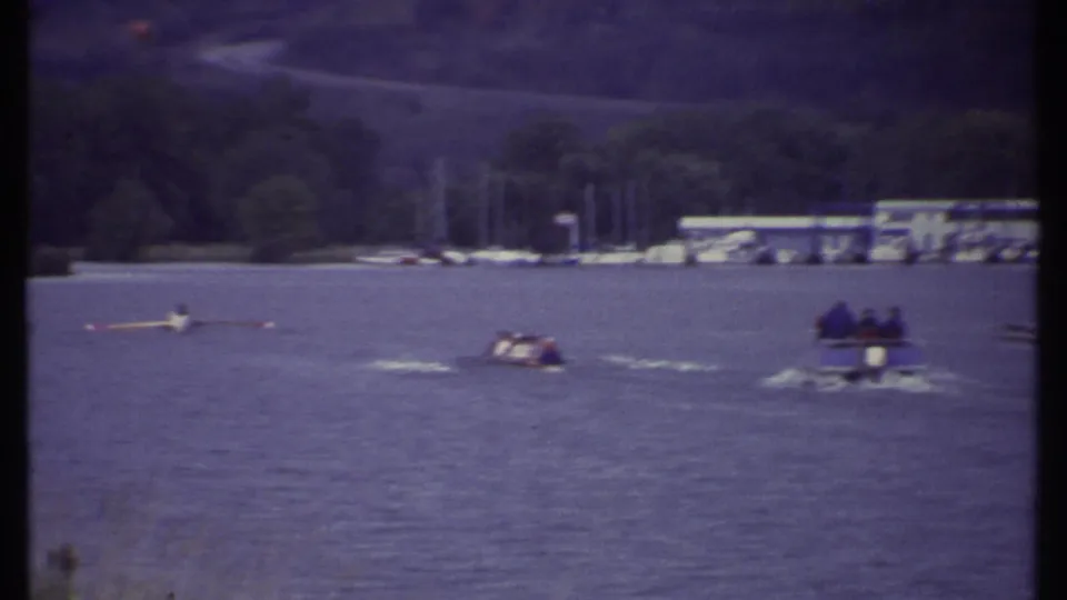 1979 Ithaca home-movie frame of scullers and a small motorboat on lake water, with a marina and trees in the background.