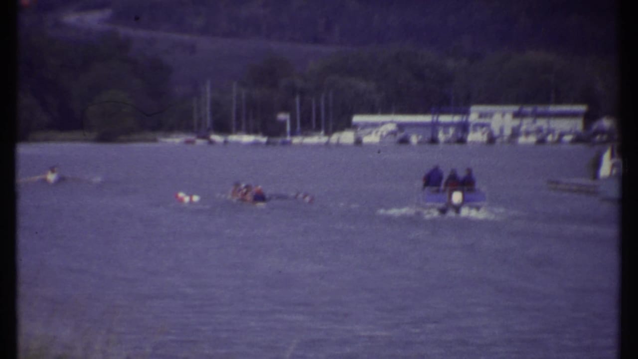 1979 Ithaca home-movie frame showing small boats on lake water, distant people, docked sailboats, and wooded hills beyond the marina.