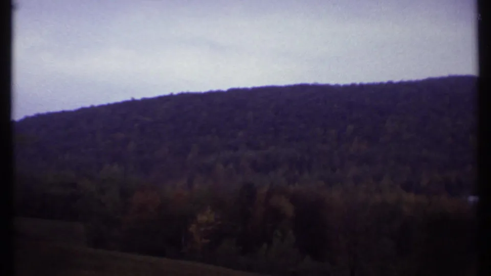 Wide 1979 Ithaca view of a wooded hillside beneath a pale overcast sky, framed by dark 8mm film borders.