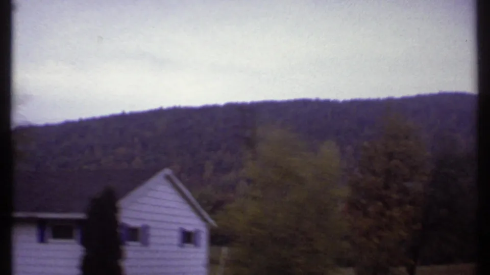 1979 Ithaca-area home-movie frame showing a white house with blue shutters near trees below a wooded hill.