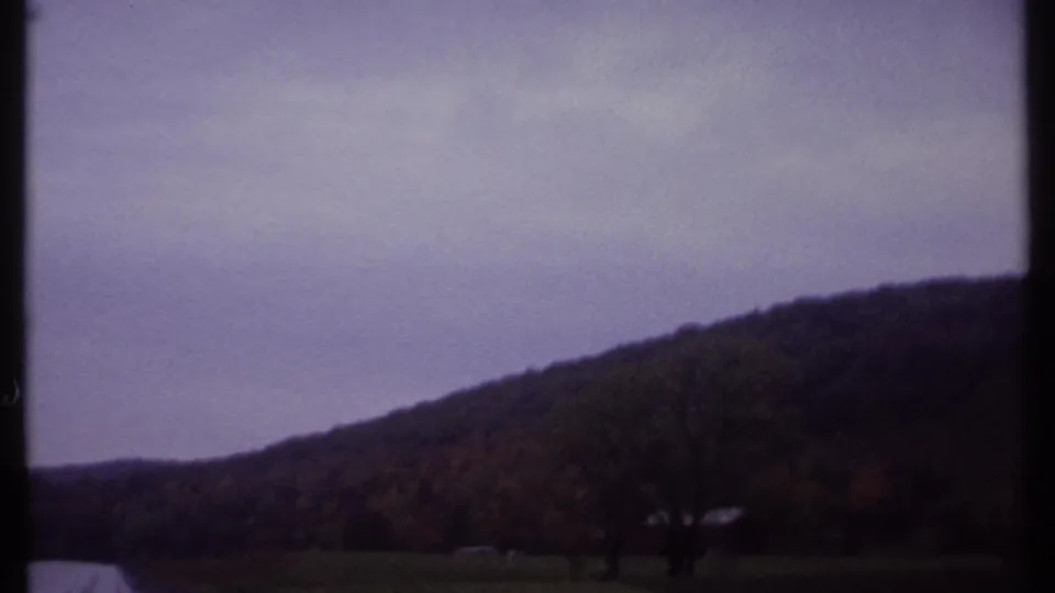 1979 Ithaca-area 8mm view of a sloping wooded ridge beyond open ground, with bare trees silhouetted under cloudy sky.
