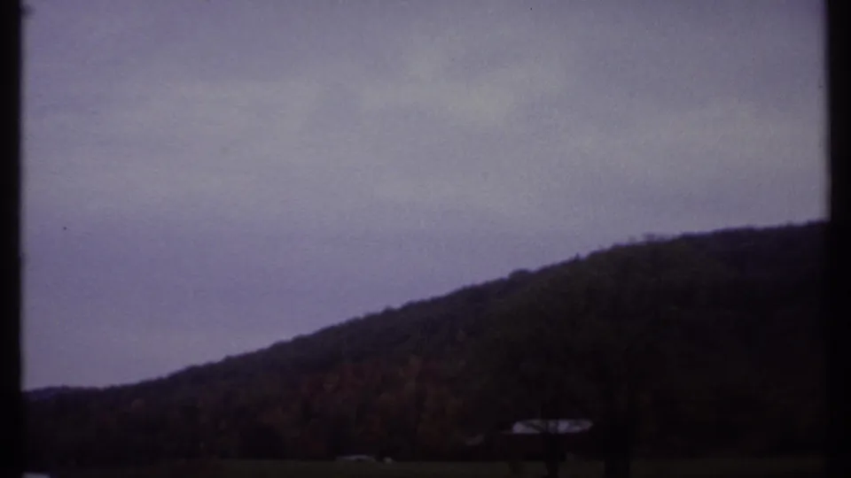 1979 Ithaca-area 8mm frame with a sloping tree-covered hill, cloudy sky, and a distant pale structure near the shoreline.