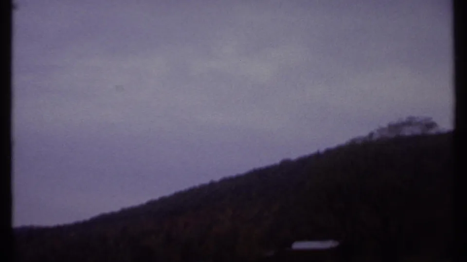 Soft 8mm frame from the 1979 Ithaca landscape clip showing a hillside, bare tree silhouette, cloudy sky, and small bright structure.