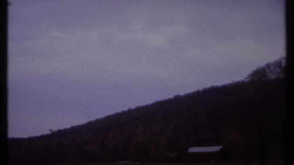 1979 8mm landscape view near Ithaca, New York, with a dark hillside, muted cloudy sky, and distant small structure.